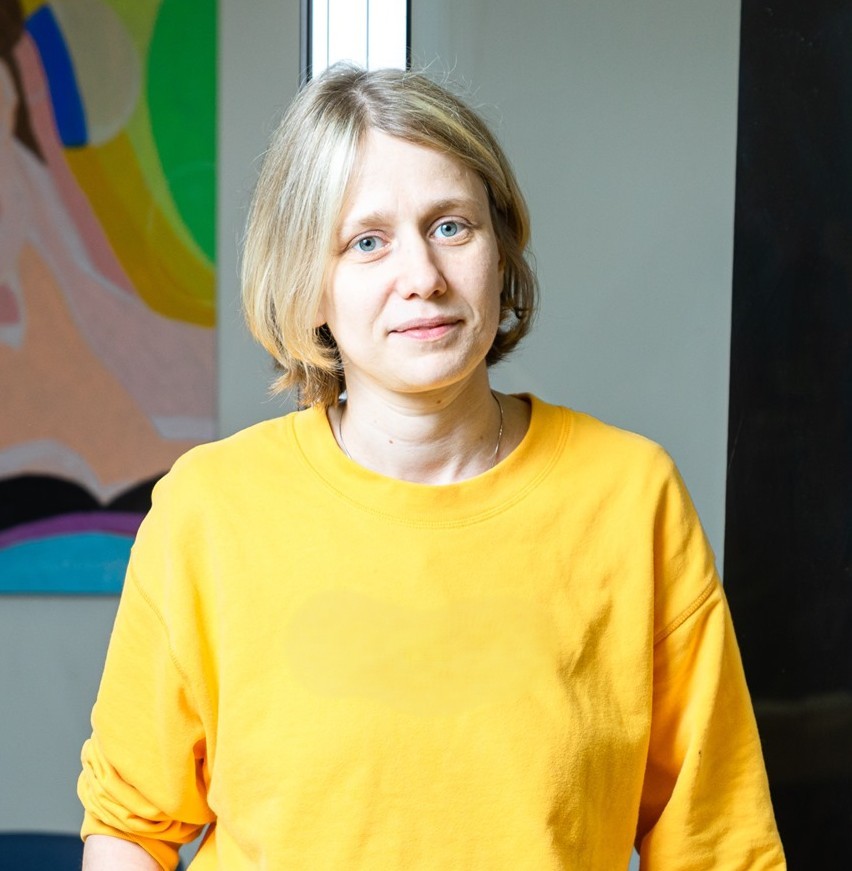 Aleksandra Pavlovska smaidīga, dzeltenā Levi’s džemperī, skatās tieši kamerā.